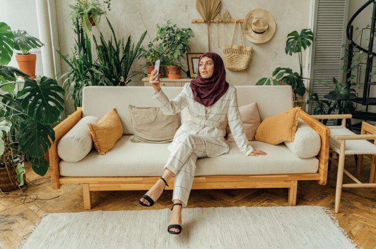 A woman wearing a hijab takes a selfie while sitting on a stylish sofa in a modern, plant-filled living room.